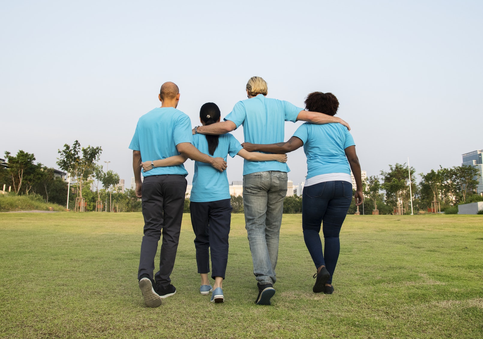 Four adults walking through the grass away from the camera with their hands around each others backs showing community. They are all wearing bright blue/teal shirts with jeans.